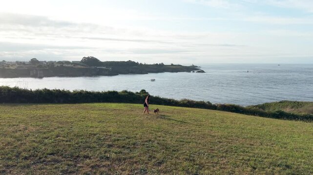 Young woman happily throwing a toy for her energetic dog in a scenic green field overlooking the ocean. Beautiful summer day of fun