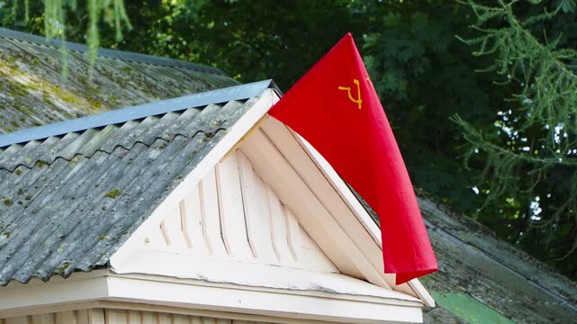 A red flag displaying the yellow hammer and sickle symbol sways in the wind on the roof gable of a vintage wooden building outdoors.