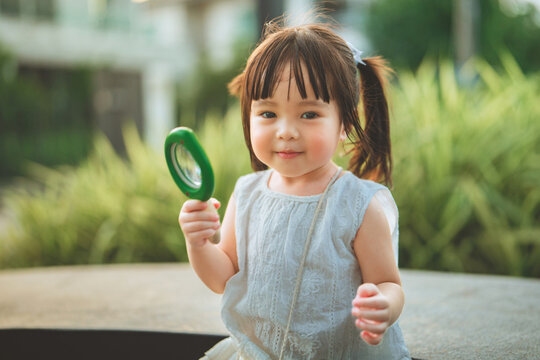 Curious toddler girl holding magnifying glass outdoors and smiling at camera, childhood exploration, discovery, nature learning, curiosity, education and playful early development concept