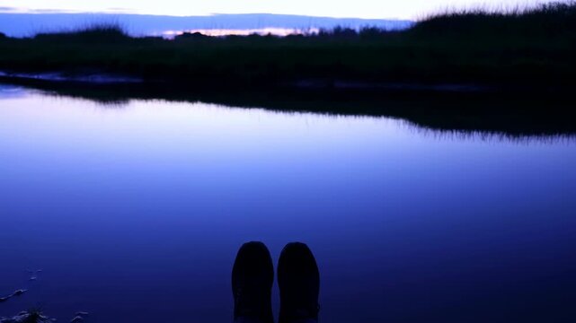 Dark shoes extend over calm reflective water at twilight, framed from a first-person viewpoint with a low grassy shoreline, soft blue tones, and a narrow band of fading light
