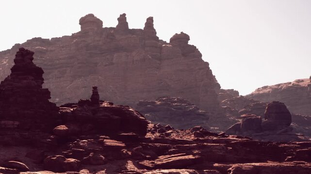 In a dry and rugged landscape, tall rock formations rise against a clear sky. The ground is rocky, with shadows cast by the structures, showing their unique shapes.