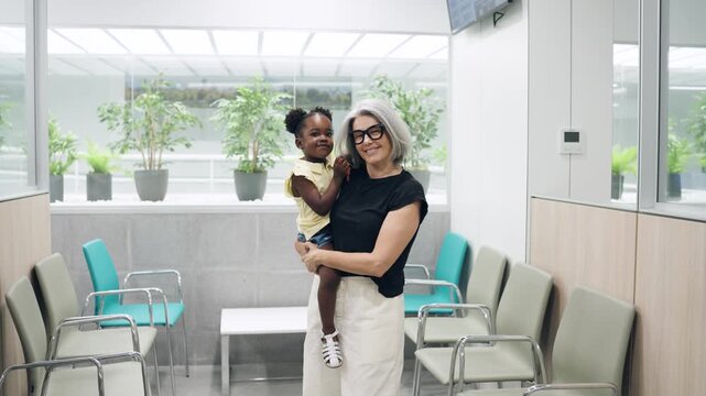 Elderly caucasian woman holding her adopted african american granddaughter in a hospital waiting room. Multiracial family waiting for a doctor