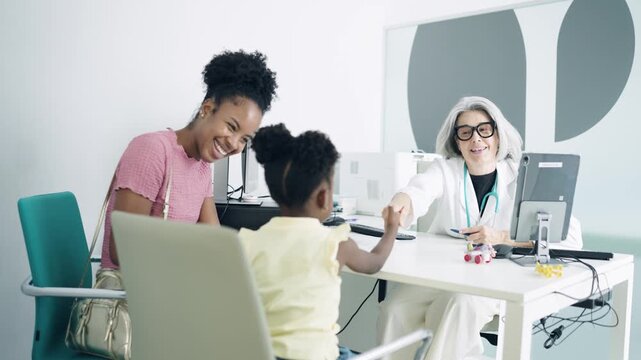 Experienced pediatrician playing and shaking hands with a little African American girl during a check up. The mother is smiling next to her daughter