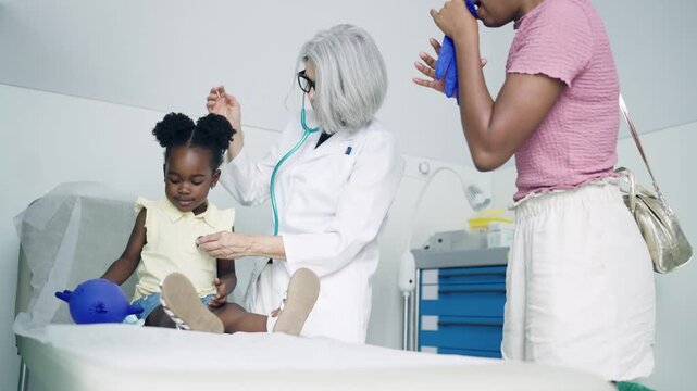 Female pediatrician examining a smiling little African girl with a stethoscope. The mother entertains her daughter by inflating a disposable glove