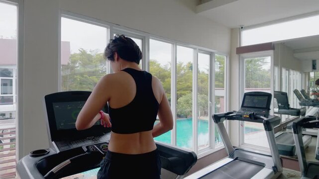 Rear view of an active Asian woman in her forties wearing a black sports bra while running on a treadmill. She exercises in a modern gym, looking out large windows during her daily cardio workout.