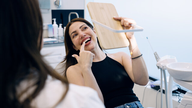 Woman smiling, seeing her teeth and pointing at them in a mirror, reflecting on dental results in a clinic