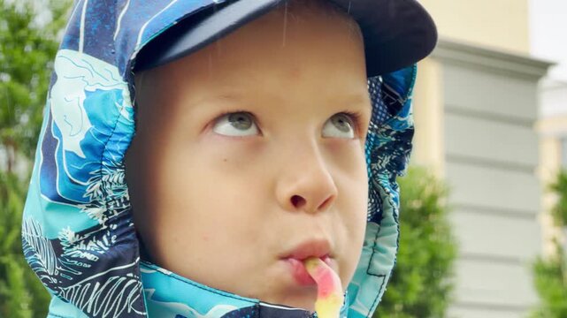A young boy in a blue jacket eats candy outdoors in the rain. The happy child enjoys his sweet treat on a gloomy street. He feels joyful, content, and pleased.
