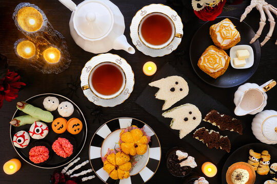 Halloween theme afternoon tea table scene against a black wood background. Spooky savory finger foods, sandwiches, pumpkin scones, and scary desserts. Top down view.
