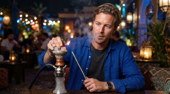 Caucasian man lighting a hookah at night. Young male using a lighter on shisha charcoal in an outdoor lounge. Evening leisure concept