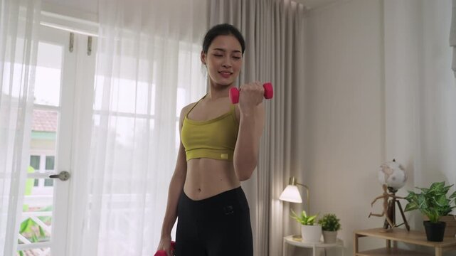 Slim beautiful Asian young woman in sportswear holding pink dumbbells to stretching physical exercise, fitness for healthcare with weight control and build muscle strength in the living room at home. 
