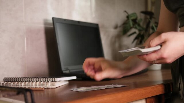 Close up side view of hands tapping stack of US dollars on wooden table and counting 100 dollar bills near laptop in home office