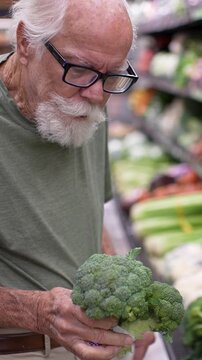 An elderly man shops for groceries in a store and examines a bunch of broccoli. He focuses on his selection in the afternoon light.