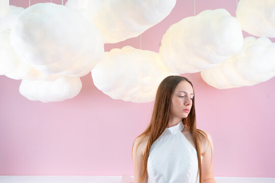 Young woman with long braided hair standing beneath oversized fluffy cloud decorations against pastel pink wall looking down with calm expression, with woman, female, young, portrait detail