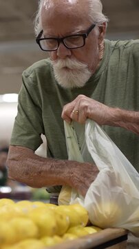 An elderly man picks lemons in a grocery store and puts them in a plastic bag while shopping for his groceries.