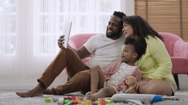 Happy Family Day concept. African American family sitting, smiling, and using a tablet for a video call while their little daughter plays with plane model, with happiness on living room floor at home.