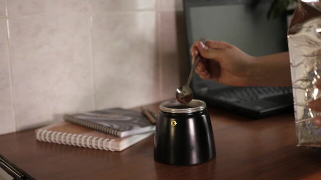 Close up side view of person putting ground coffee into moka pot filter with spoon and screwing top part on wooden table in home office