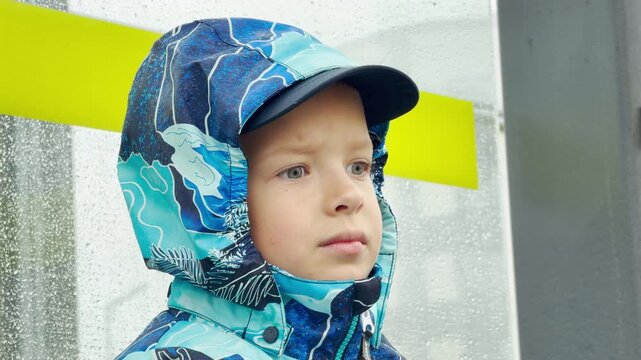 A young boy in a blue hooded jacket looks thoughtfully forward. He waits inside a rainy bus shelter during a storm. This quiet, melancholic environment feels calm yet lonely.