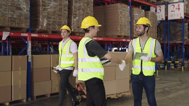 Teamwork of men warehouse workers in yellow helmets holding a tablet, working, talking, discussing, planning, inventory, moving cardboard boxes by hand pallet truck in logistics distribution factory. 