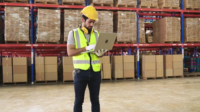Young man warehouse worker in uniform wearing a yellow safety helmet, standing, holding, and using a laptop computer for analysis while checking inventory stock in a logistics distribution factory. 