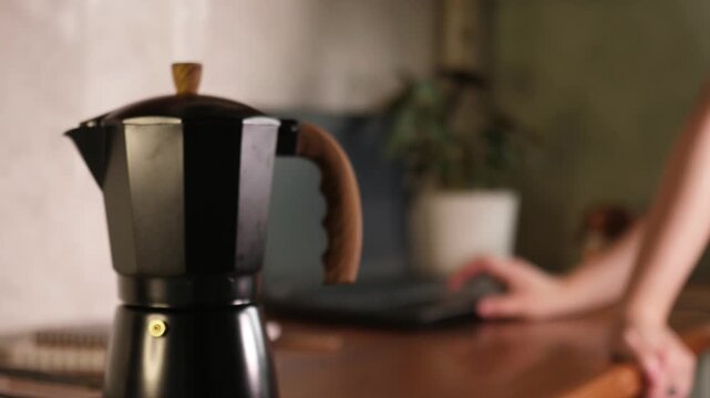 Selective focus close up of moka pot coffee maker on wooden table with blurred woman working on laptop in background at home office