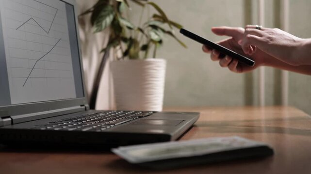 Close up side view of person hands using smartphone screen next to laptop with financial growth chart and cash stack on wooden table
