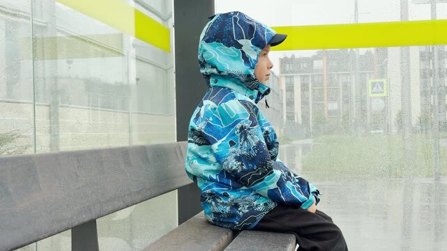 A young boy in a blue raincoat sits on a wooden bench inside a bus shelter. Raindrops cover the glass. This rainy weather creates a lonely, melancholic, and gloomy mood.