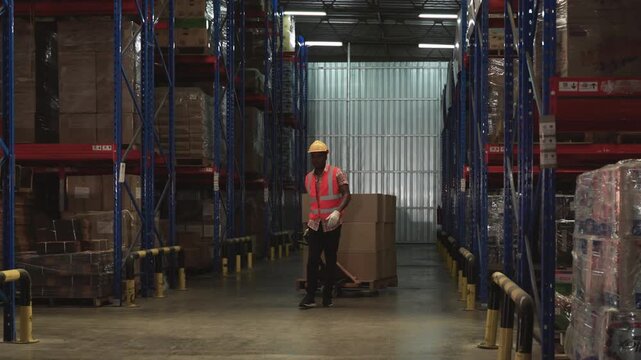 African American male warehouse worker in orange vest uniform wearing yellow safety helmet using hand pallet truck for moving cardboard boxes of cargo from shelves in logistics distribution factory. 