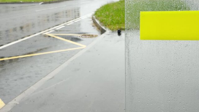 A wet glass panel with a yellow stripe stands near a rainy asphalt street. Water droplets cover the surface overlooking an empty road. The dreary environment feels quiet and melancholic.
