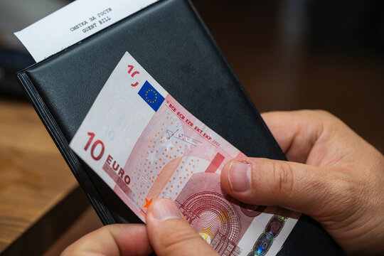 A person holds a black bill folder containing a ten-euro banknote and a guest receipt.