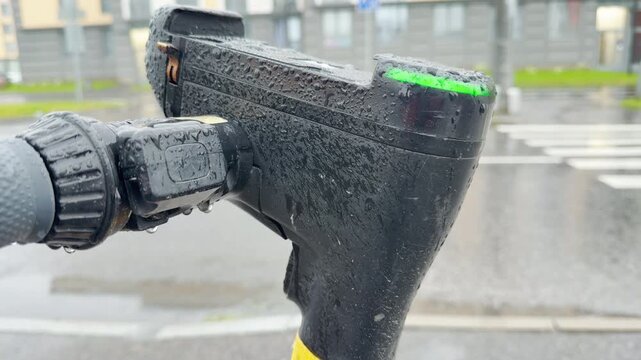 A wet electric scooter handlebar stands in the rain. Water droplets cover the plastic surface near an urban street. This gloomy, melancholic environment feels cold and lonely.