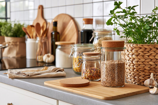 Cozy kitchen scene with wooden cutting board and jars. Fresh herbs sit beside pantry items and cooking tools. Natural light highlights tidy, rustic countertop setup. Organic textures blend warmth