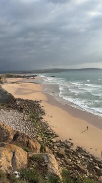 The returning Atlantic tide sweeps across a wild sandy beach beneath a heavy stormy sky, while fading sunset light breaks through and turns the ocean water shades of grey and gold in Cantabria, Spain.