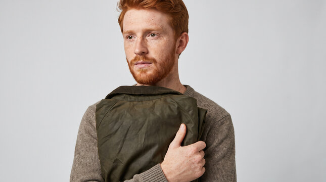 Young freckled redheaded man holding folded jacket against plain light studio background. Concept of raincoat, seamless, backdrop, freckles, studio portrait. Copy space.
