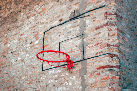 Red metal basketball rim mounted on an old weathered brick wall with painted backboard outlines.