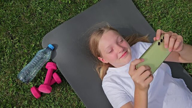 Girl in white t‑shirt smiling, holding smartphone, lying on grey mat outdoors. Pink dumbbells and plastic water bottle on grass. Natural daylight, relaxed atmosphere, fitness break.