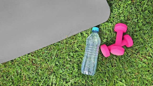 Close‑up outdoor shot showing a dark grey fitness mat partially in frame on the left, its smooth textured surface angled diagonally. On green grass in the foreground &mdash; a transparent plastic water bott