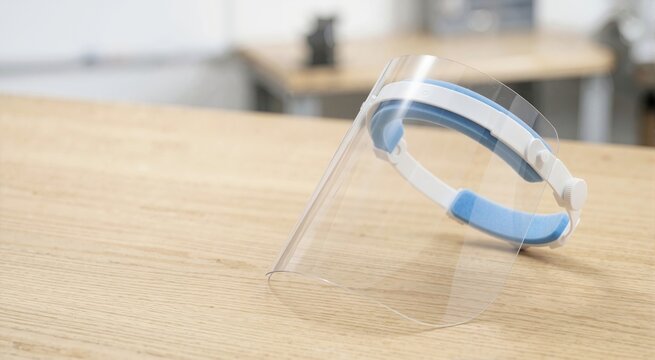 Clear plastic protective face shield resting on a wooden table. Personal protective equipment with white headband and blue foam padding