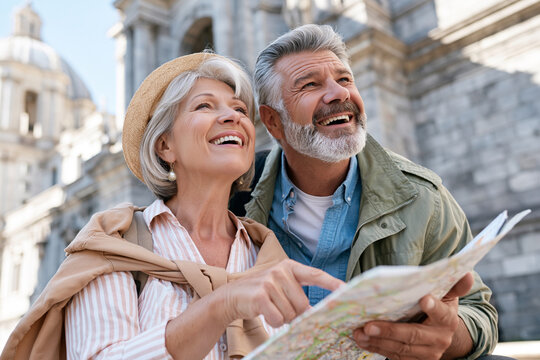 Senior couple laughs, exploring city streets together. They point at map, sharing joy of discovery. Grand architecture looms behind them, sunlit. Warm tones, candid moment, travel adventure vibe