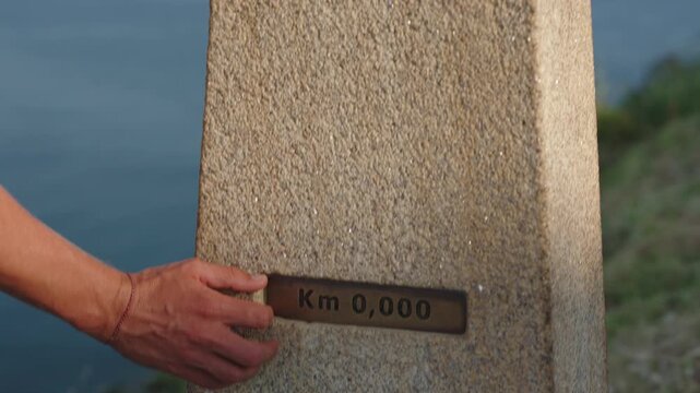 Close-up shot of a hand touching a stone marker inscribed with "Km 0,000," on Santiago way symbolizing the starting point of a journey or pilgrimage.