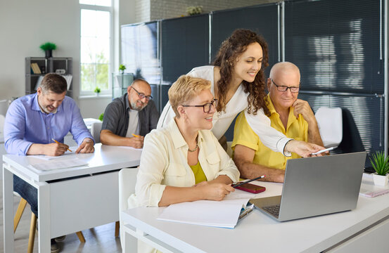 Adults seniors students with instructor during computer training. In a modern classroom they learn at a laptop, build digital skills and confidence. Concept: lifelong learning and upskilling.