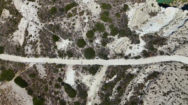 Aerial drone view looking down over dirt trails on the Greek island of Milos. Two people can be seen walking the pathway in Greece for exercise outside in the dry landscape. 