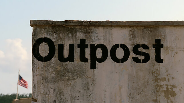 A weathered concrete wall with black painted text reading outpost under a clear sky with a distant national flag visible in the background