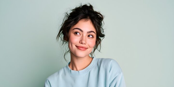 Playful young woman looking away with cheeky smile and dimples, cheerful female portrait against teal background, casual studio lifestyle photography