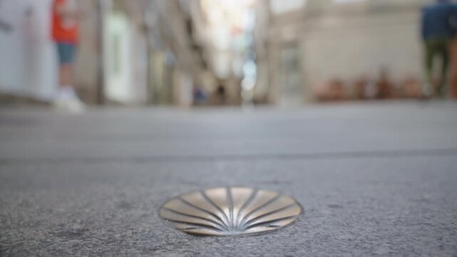 Close-up, low-angle shot of a bronze scallop shell marker embedded in a stone walkway, with the blurred feet of a person walking past in the background.