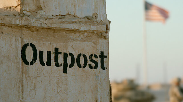 A close up view of an outpost sign on a concrete wall in a desert base setting with a flag and a soldier in the blurry background view