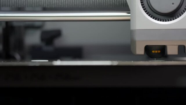 Modern device moves precisely over a flat surface. Gray casing holds a small black component. Yellow LED lights blink in sync with motion. Tool performs detailed work with mechanical accuracy
