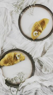 croissant,golden brown pastry,baked goods,flaky pastry,layered crust,browned surface,round plate,dark rim,white flower,yellow center,tiny blossoms,floral stem,branching stem,white fabric,draped fabric