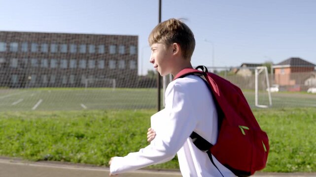Young boy walks toward school, red backpack prominent. He pauses near a pole, looking at building. Sunlight glints off his hair, soft, natural light. He seems calm, ready for the day ahead