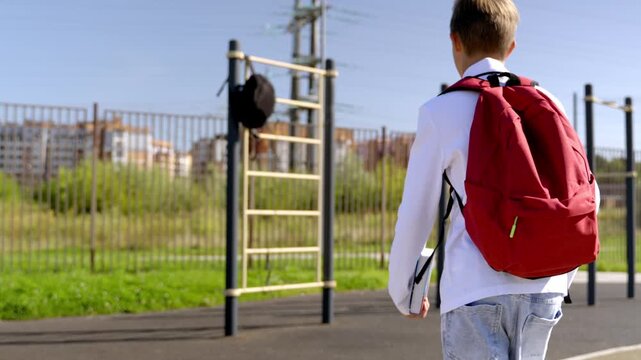 Young boy walks toward school, red backpack prominent. He pauses near a pole, looking at building. Sunlight glints off his hair, soft, natural light. He seems calm, ready for the day ahead