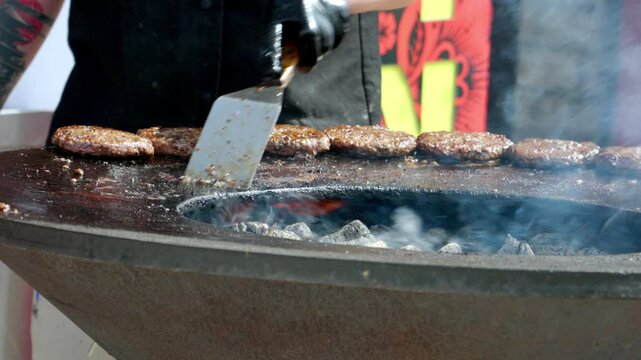 Smoke rises from sizzling burger patties over hot coals. Chef flips each patty with a spatula, expertly. Flames glow beneath a large, round, charcoal grill. The scene suggests outdoor street food